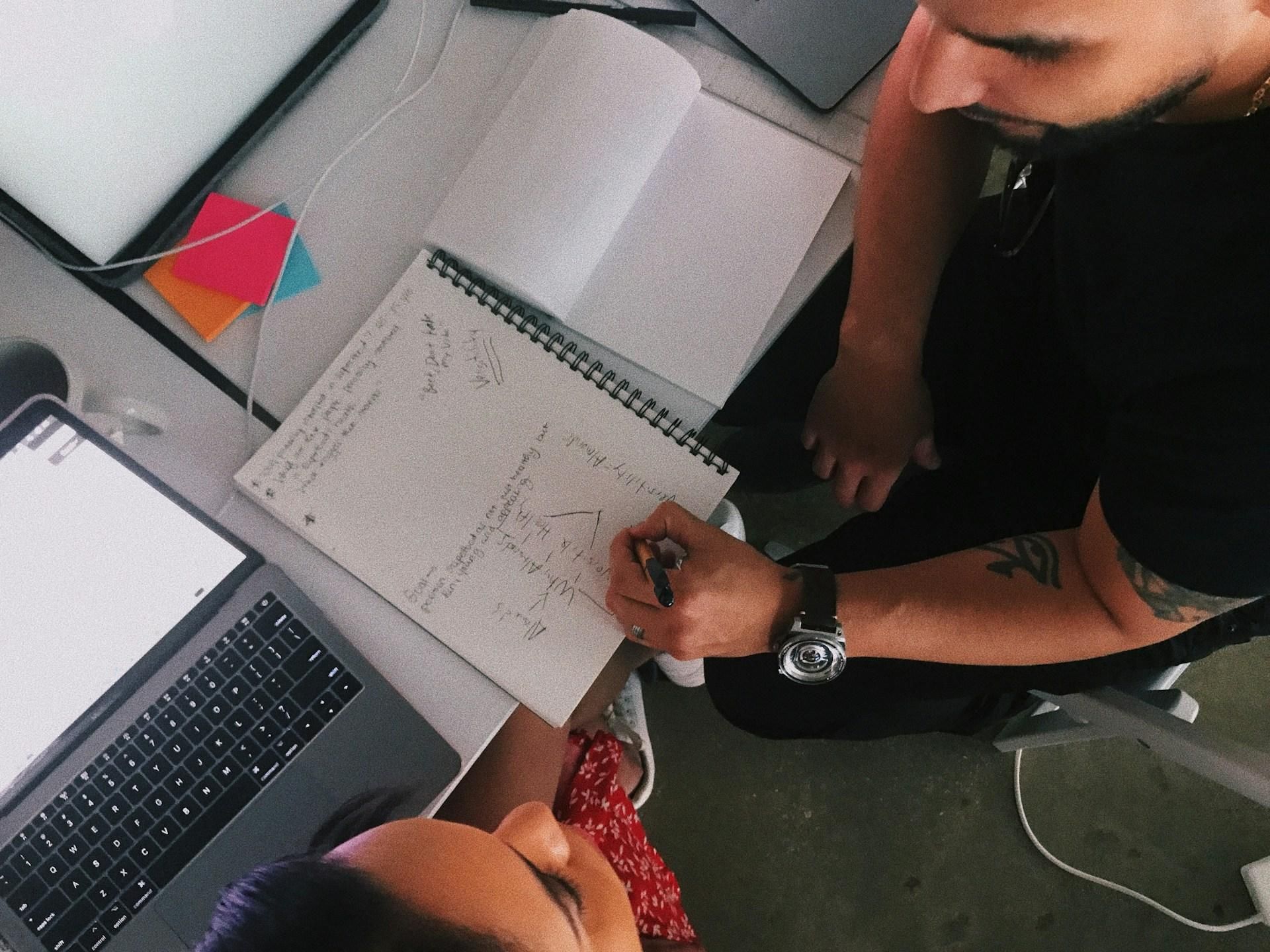 Overhead view of two students sketching notes and diagrams in a notebook beside a laptop during a project